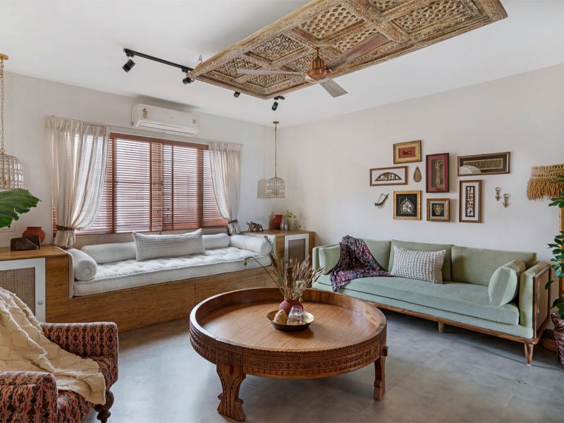 Cosy living room with a carved wooden round coffee table, a sage green sofa, a built-in wooden window seat with white cushions, framed art on the wall, and decorative pendant lights, blending modern and rustic styles.