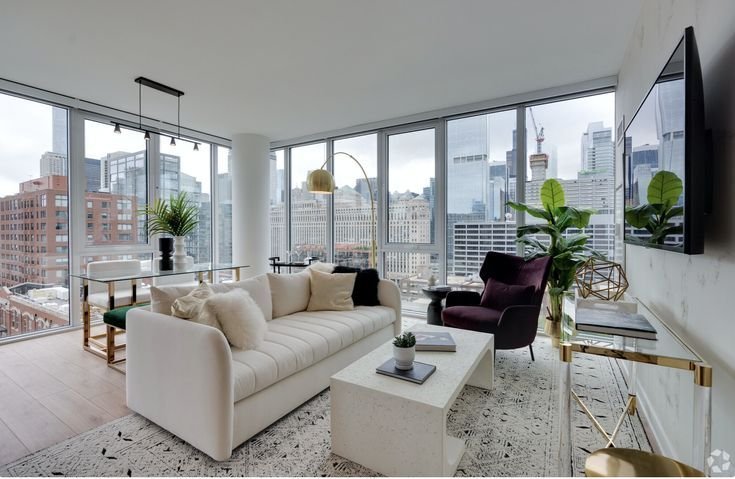 Modern apartment living room with a city view from floor-to-ceiling windows. Features include a white couch, a purple armchair, and a glass dining table.