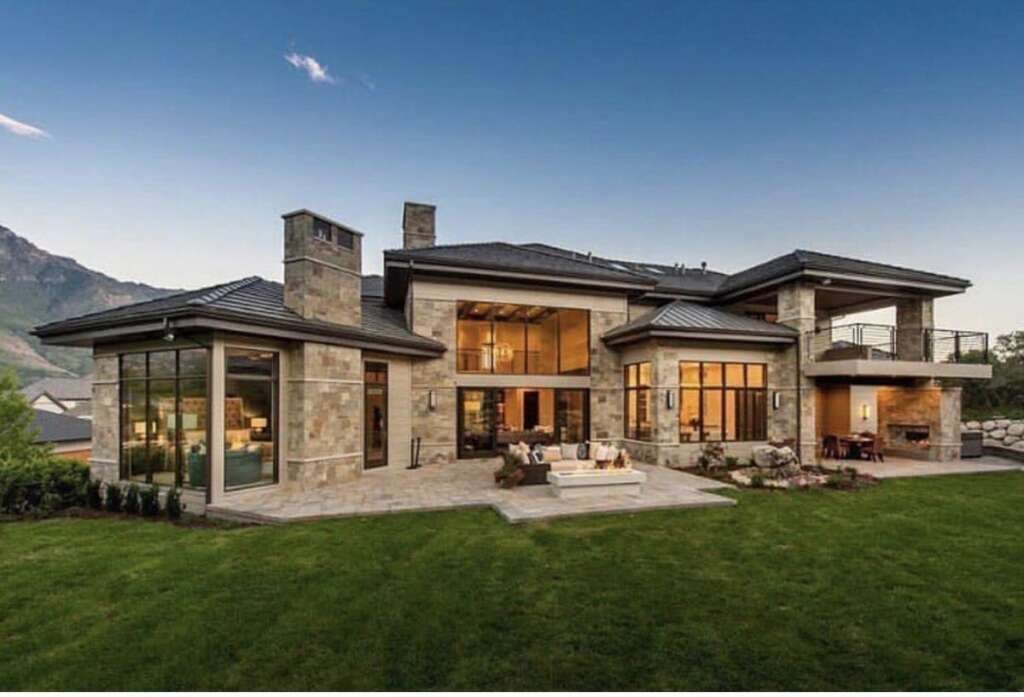 A modern farmhouse with a stone facade and expansive glass windows, featuring a patio with a fire pit and a mountain backdrop.