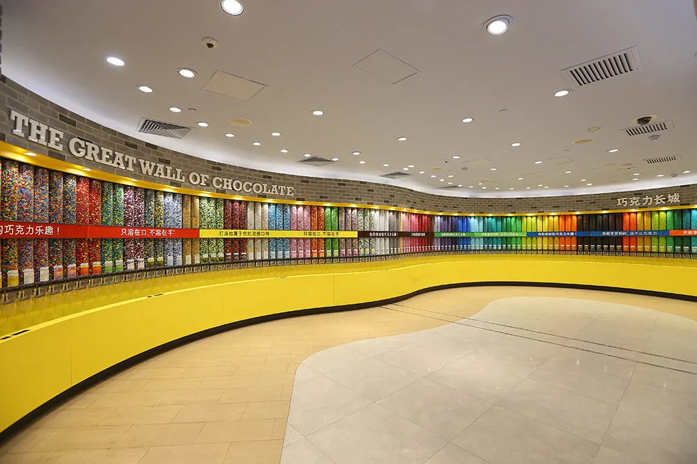 A colorful interior shot of the M&M's World store in Shanghai, showing "The Great Wall of Chocolate," a long, curved wall filled with a rainbow of M&M's candies.