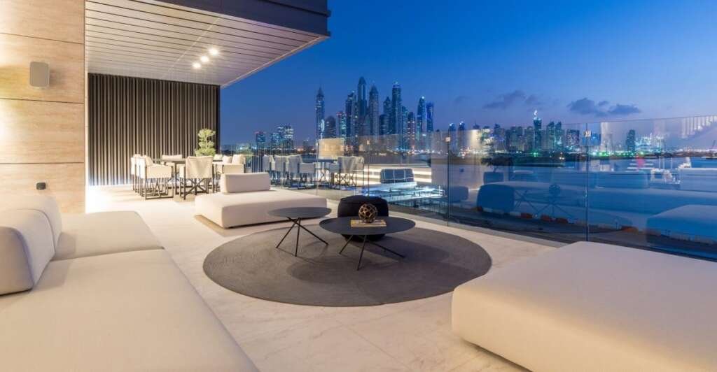 A luxurious modern penthouse terrace at dusk, featuring sleek white outdoor furniture and a stunning panoramic view of a lit-up city skyline across the water.