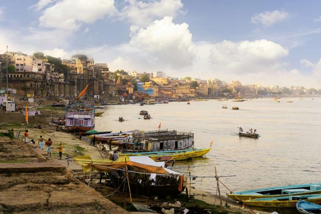Boats docked along the ghats of the Ganges River in Varanasi, India, with historic buildings and people near the water.