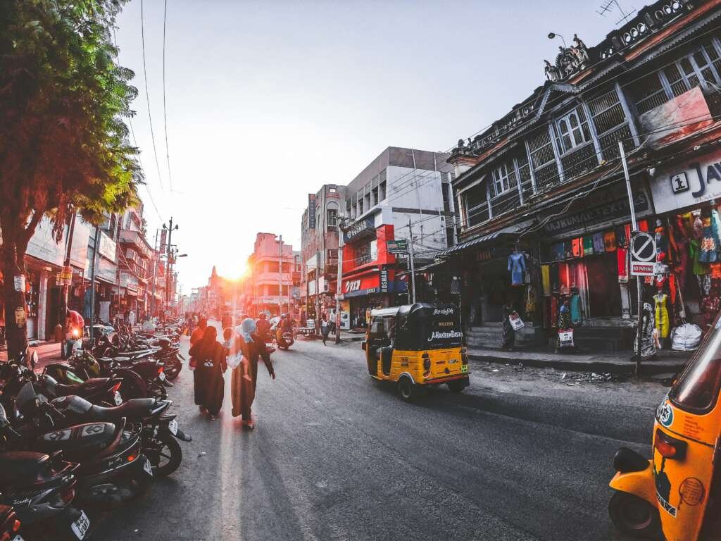 Street scene in India at sunset with people walking, motorcycles parked, shops on both sides, and auto rickshaws.