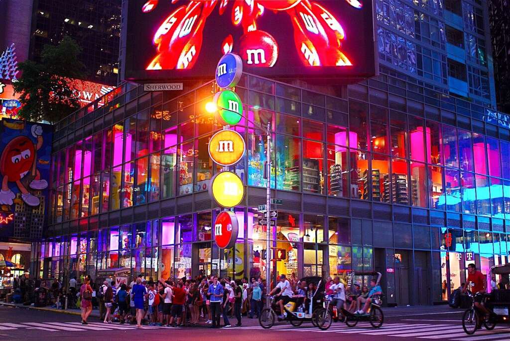A bright night view of the M&M's World store in Times Square, NYC, featuring vibrant pink and blue lights, a large video screen, and a busy crowd.