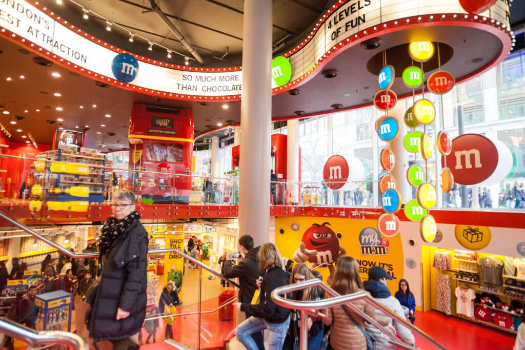 A bustling interior shot of the M&M's World store in London, with shoppers on multiple levels, vibrant red decor, and a fun red double-decker bus display.