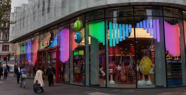 An exterior shot of the M&M's World store in London, showcasing its large glass windows with vibrant, multi-colored light displays and iconic M&M character statues. 