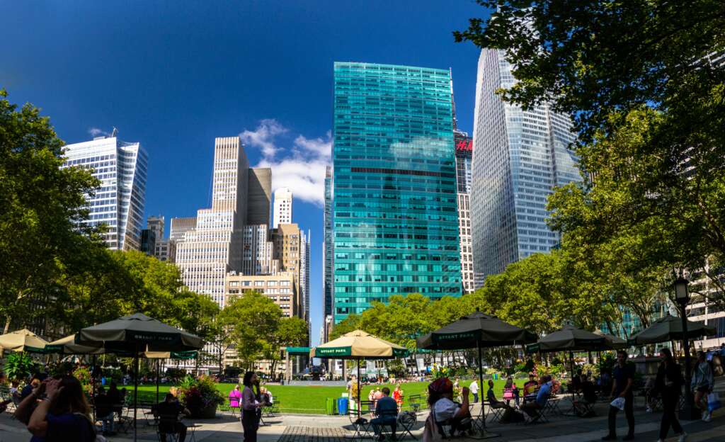 Bryant Park in New York City with people relaxing under umbrellas, green trees, and tall skyscrapers in the background.
