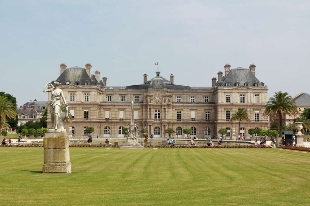 Luxembourg Palace in Paris with manicured lawns, statues, palm trees, and people walking in the Luxembourg Gardens.